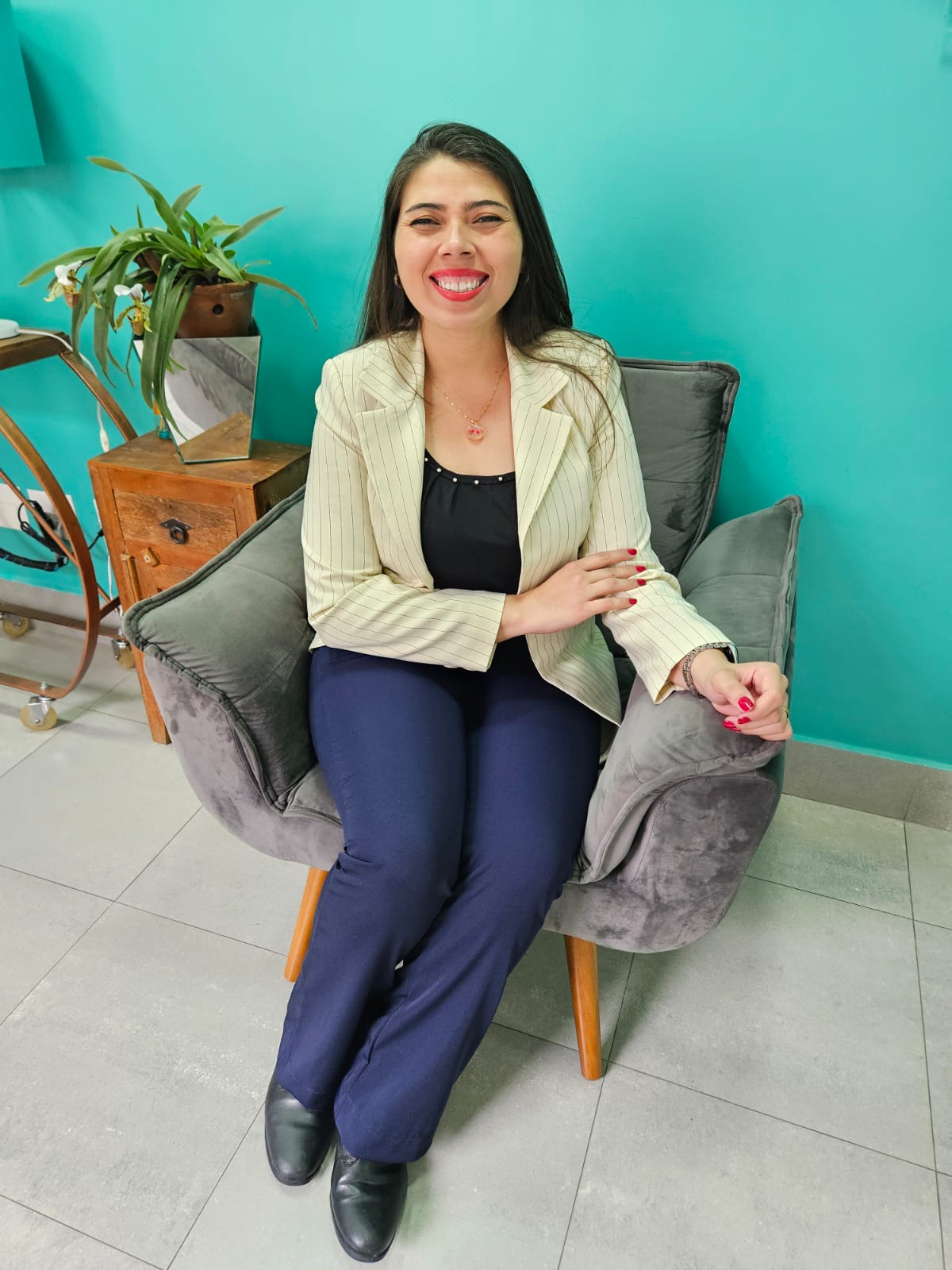 Psicóloga Luana sentada em uma portona cinza vazo de planta fundo da parede verde agua